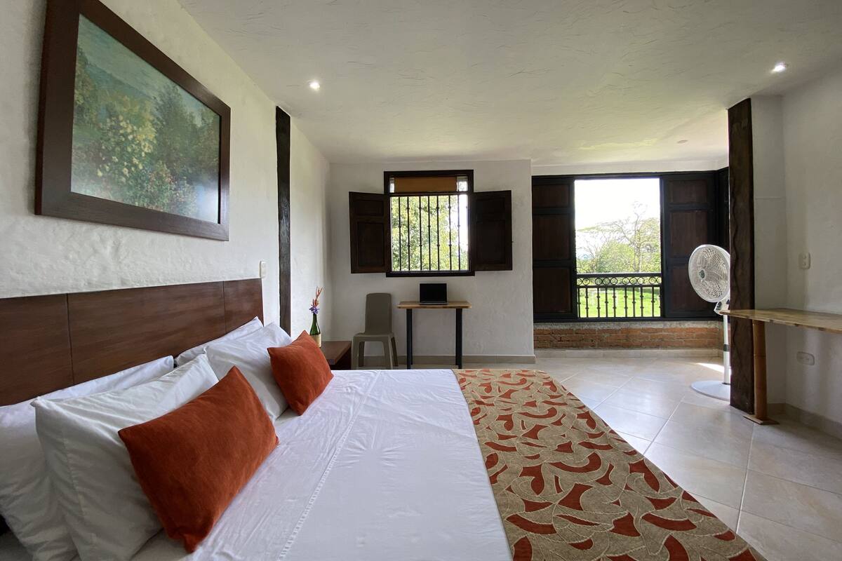 A hotel room with a large bed, a desk, a chair, a TV, and a balcony with a view of greenery.
