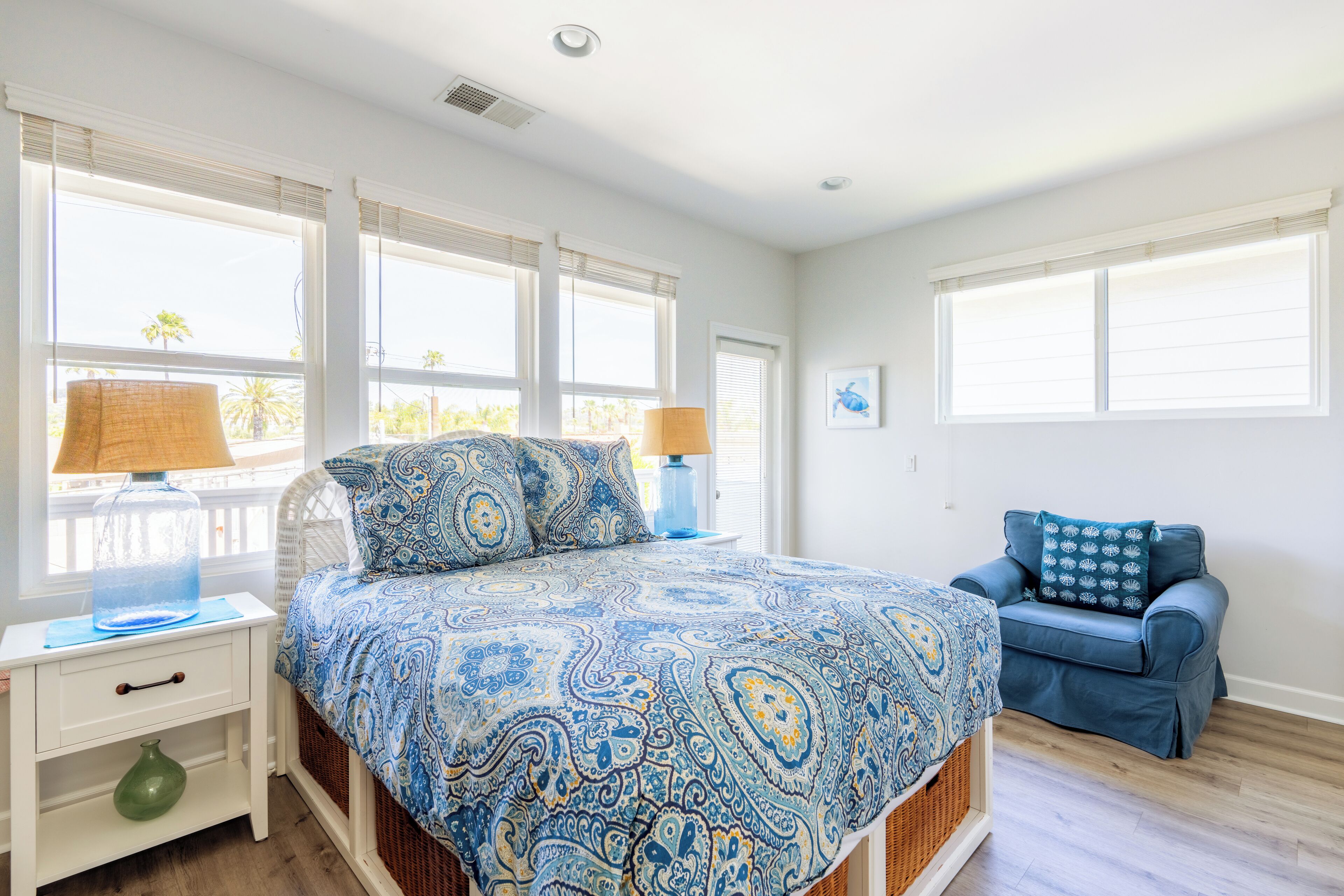 A bedroom with a large bed, two bedside tables, a blue patterned bedspread, a blue sofa, and a window with blinds.