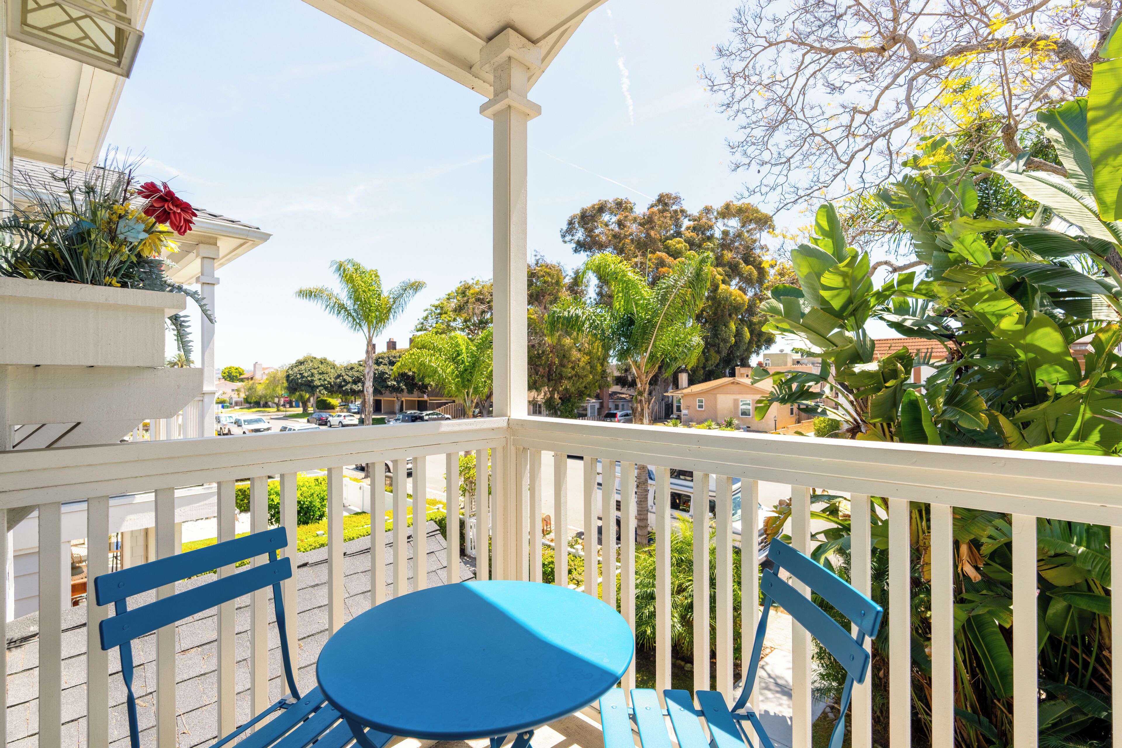 Un balcon avec une table et des chaises bleues, entouré d’une végétation luxuriante et offrant une vue sur la rue.