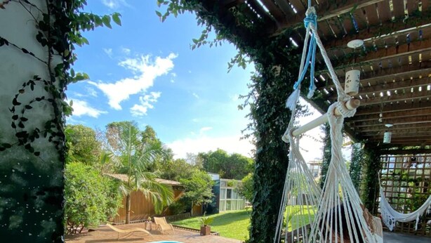 A hammock chair suspended from a wooden pergola, overlooking a swimming pool with a wooden deck and a lush garden. - Guarda Beach House - Hostel (Palhoça)