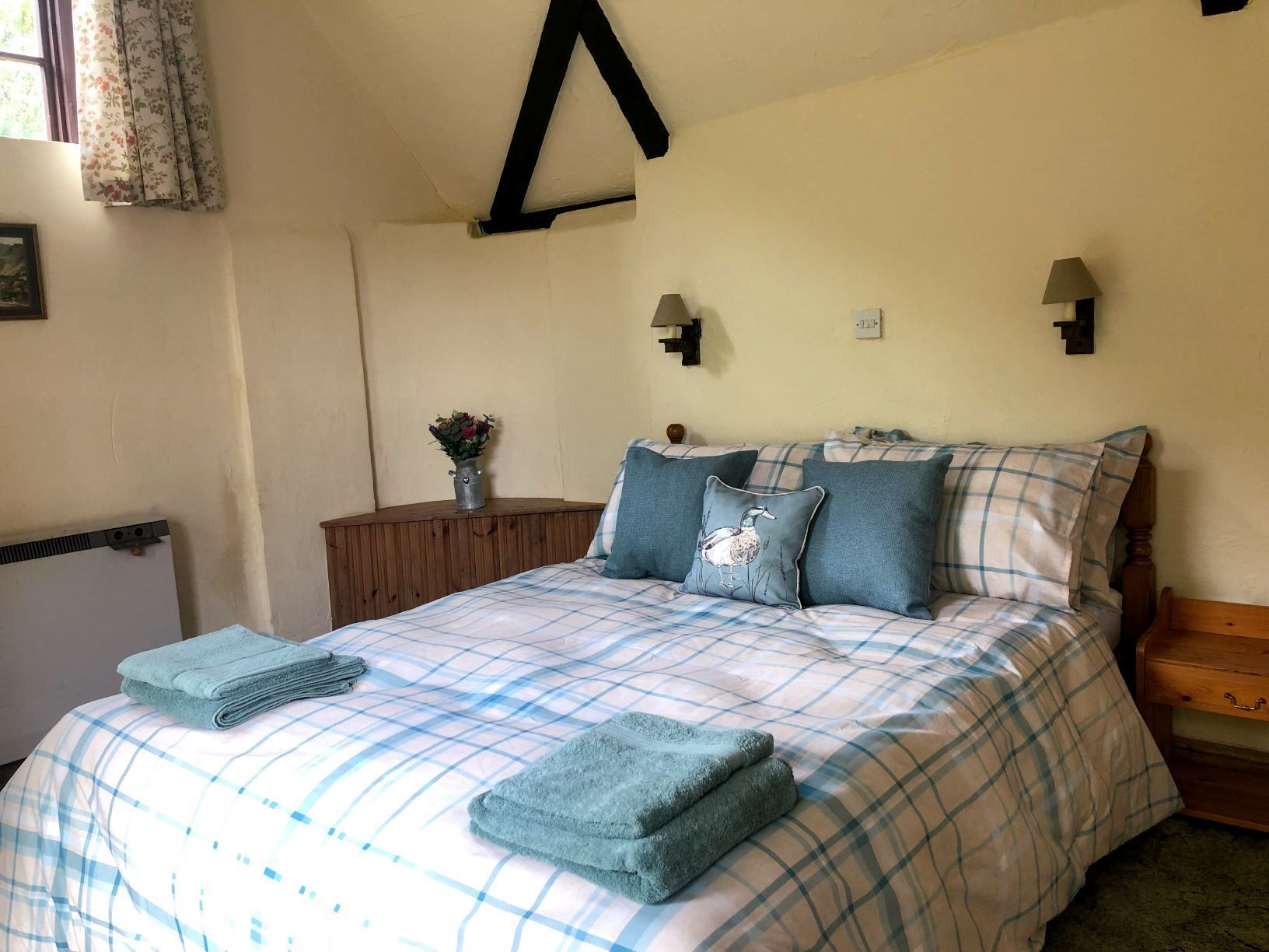 A bedroom with a bed, pillows, a nightstand, a wall-mounted lamp, and a small table with a plant.