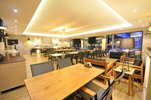 Un comedor moderno con mesas y sillas de madera, una cocina al fondo y ventanas amplias que ofrecen vistas de la ciudad.