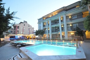 Un área de piscina con sillones de descanso, un bar y un edificio de varios pisos con balcones.