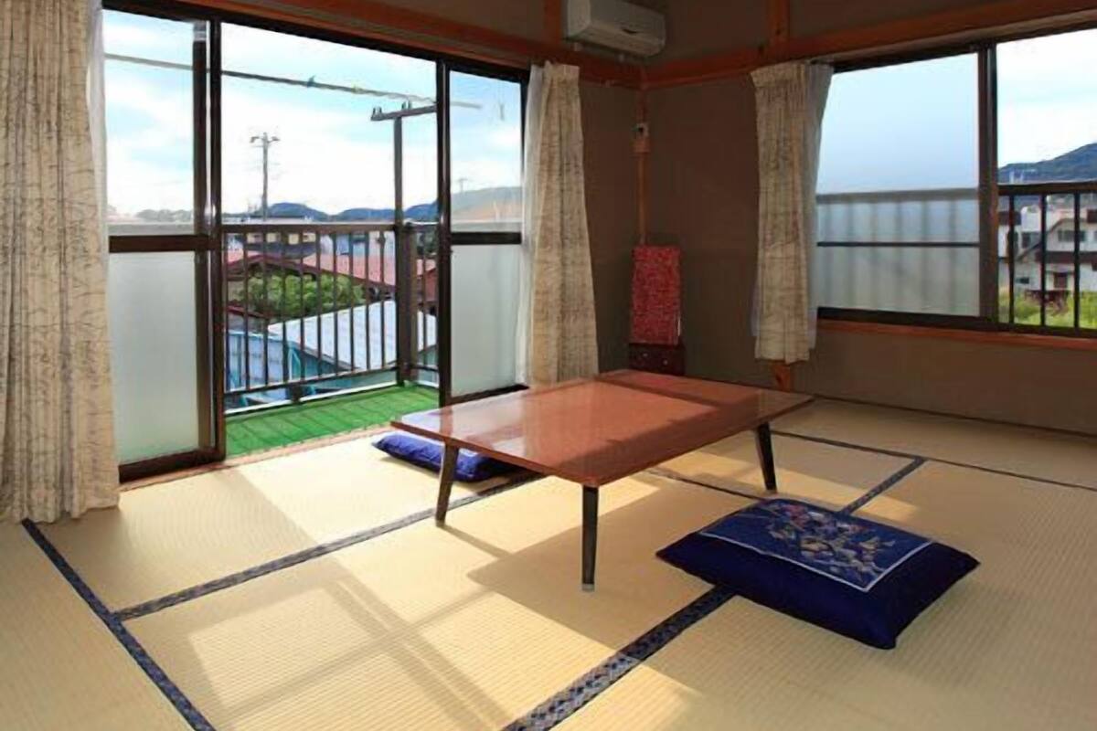 A tatami mat room with a low table, sliding glass doors, and a view of a green landscape.