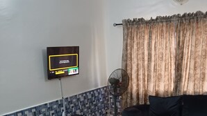 Un salon avec un canapé bleu, un téléviseur fixé au mur, un ventilateur sur pied et un rideau à motifs.