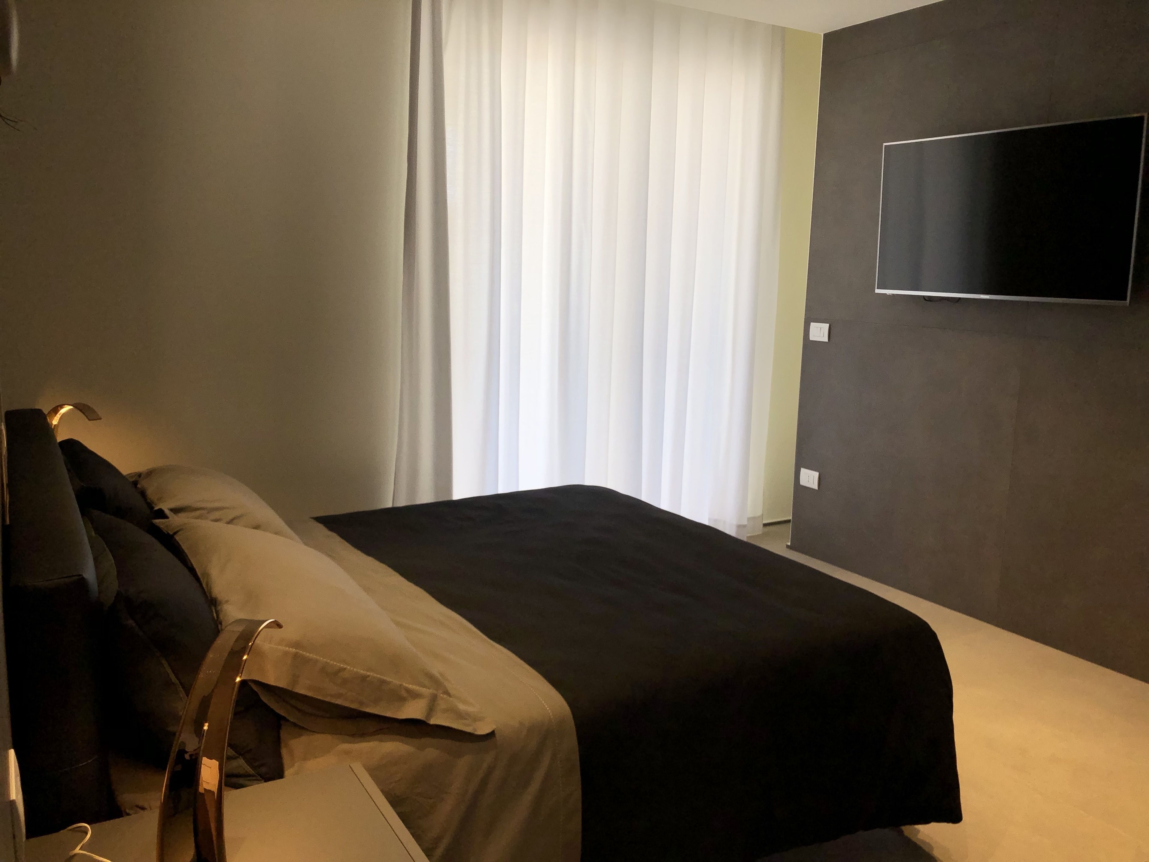 Una habitación de hotel moderna con una cama, un televisor de pantalla plana en la pared y un ventanal con cortinas traslúcidas.
