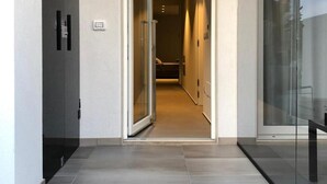 Un pasillo moderno con una puerta de cristal que da a una habitación. El pasillo tiene suelo de baldosas y un interruptor de luz en la pared.