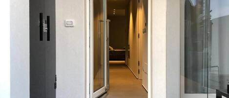 A modern hallway with a glass door leading to a room. The hallway has a tiled floor and a light switch on the wall.
