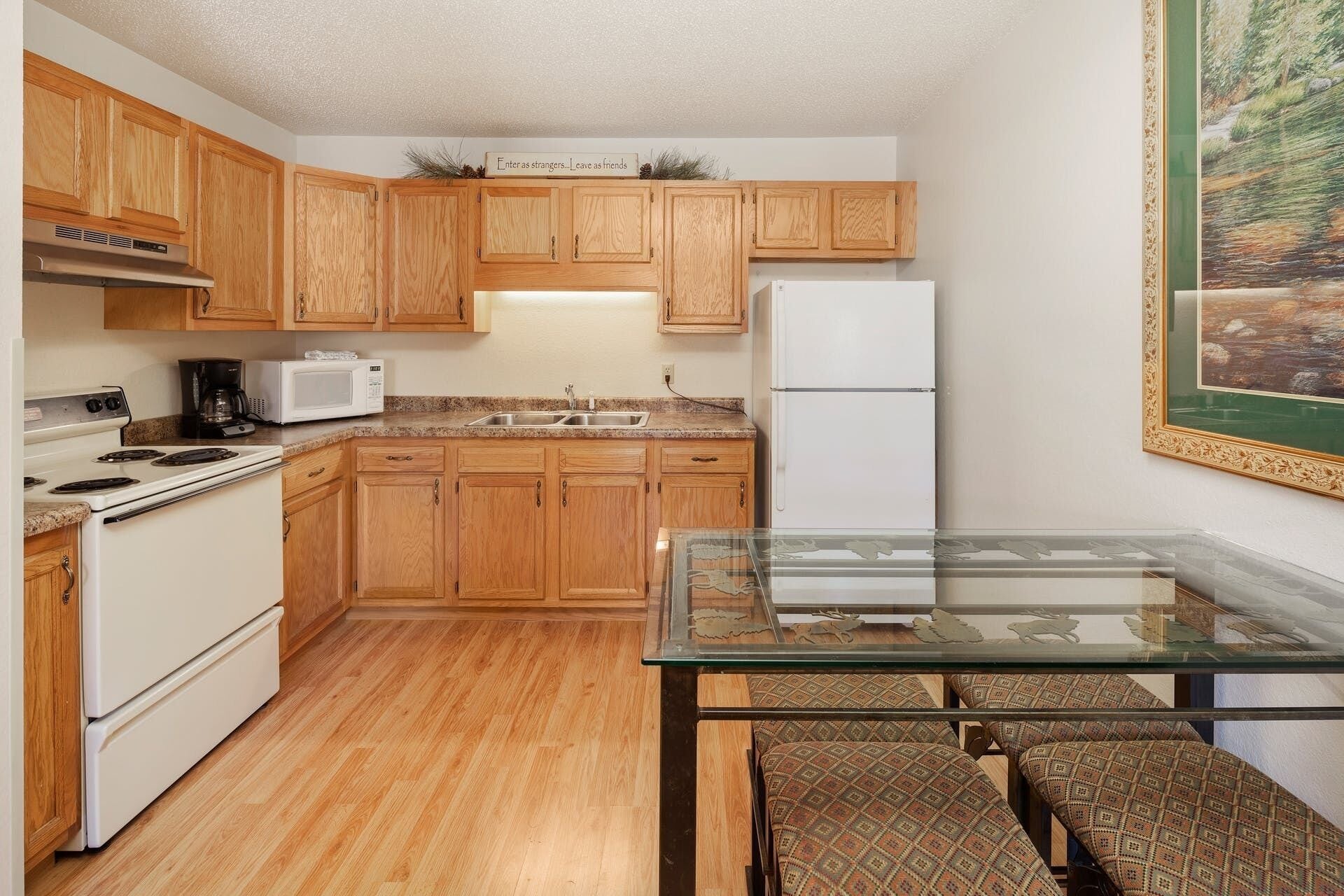 A compact kitchen with wooden cabinets, a white stove, a microwave, and a refrigerator. There's a glass table with chairs and a painting on the wall.