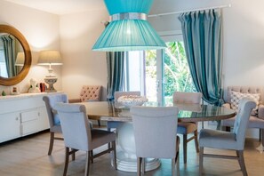 A dining area with a round table, chairs, a blue pendant light, and a view of greenery outside.