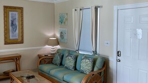 Ein Wohnzimmer mit Sofa, Couchtisch und Korbstühlen. Eine Tür, ein Deckenventilator und Wandbilder sind ebenfalls zu sehen.