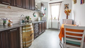 Cocina con armarios de madera oscura, encimera blanca, un barril de madera y un comedor con una silla blanca y mantel naranja.