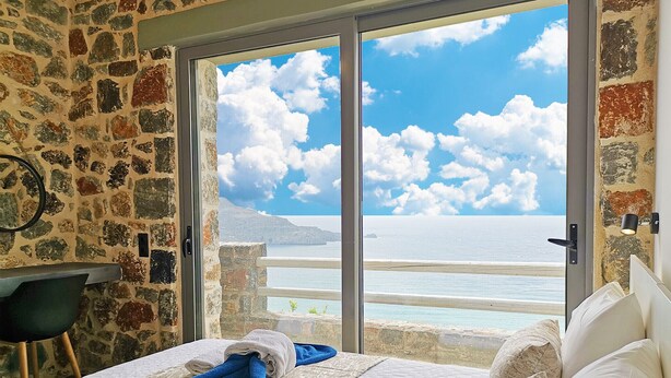 Ein Zimmer mit einer Steinsandsteinwand, einem Bett mit weißen und blauen Handtüchern und Blick auf Meer und Himmel durch eine Glastür.