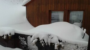 Una cabaña de madera con fachada nevada y una ventana con cortina.