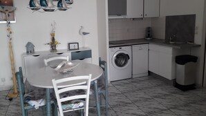 Een compacte keuken met een wasmachine, een eettafel met stoelen, en decoratieve wanddecoratie van een zeilboot.