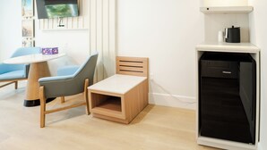 Une pièce de taille modeste comprenant un lit, une petite table, une chaise, un téléviseur fixé au mur et un coin cuisine compact avec un lave-vaisselle.