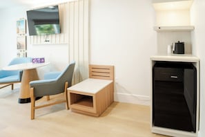 A compact room with a bed, a small table, a chair, a television mounted on the wall, and a compact kitchen area with a dishwasher. - The Belmont Shore Inn (Long Beach)