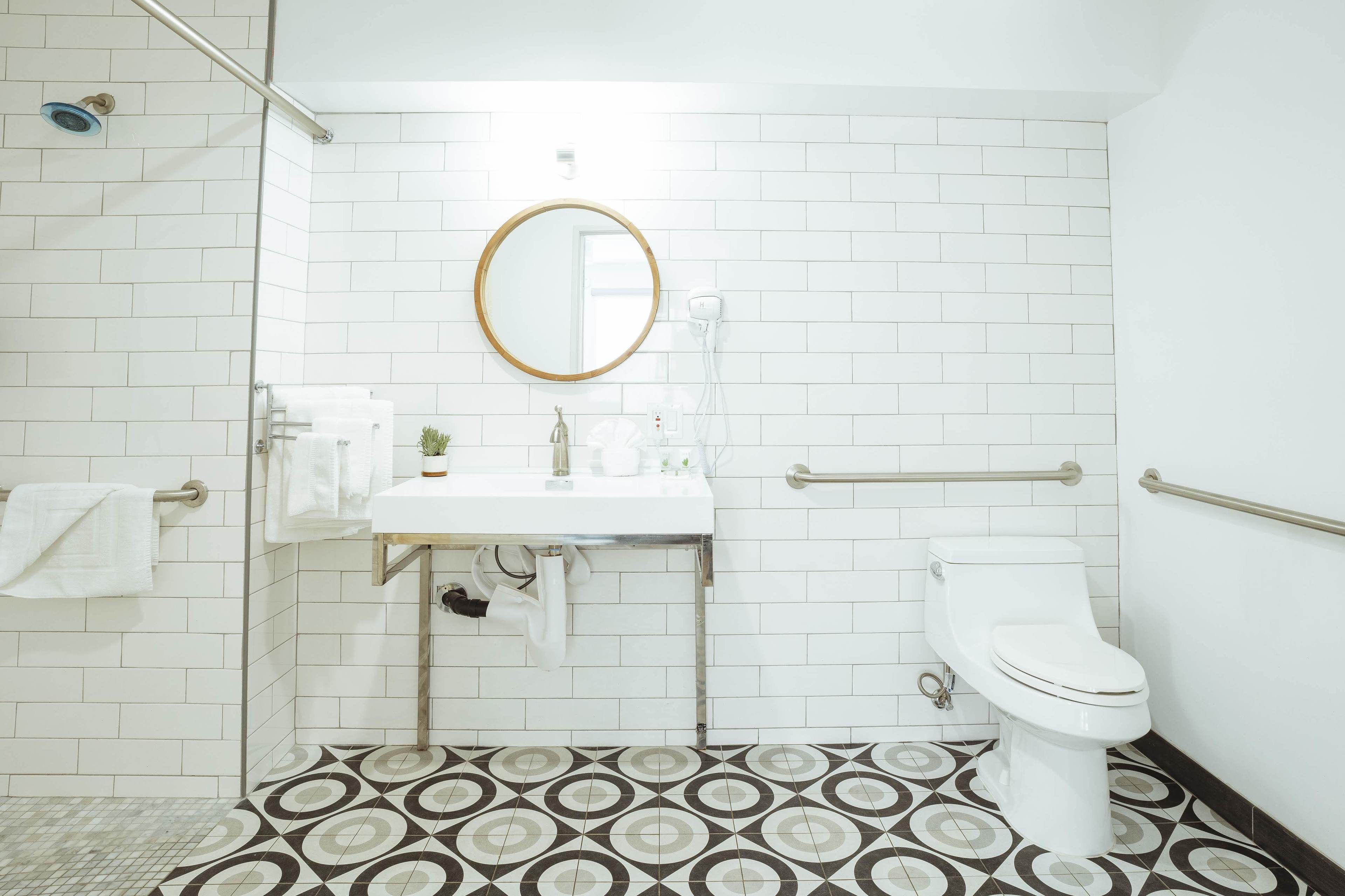 A bathroom with a white tiled wall, a round mirror, a white sink, a toilet, and a patterned floor.