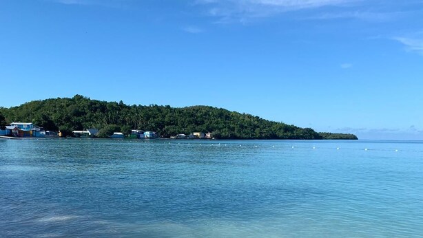 Una playa con aguas cristalinas y poco profundas, una orilla arenosa y una pequeña isla con edificios al fondo.
