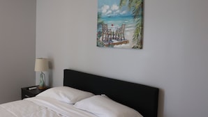 Un dormitorio con cama, una mesita de noche con lámpara y un cuadro con motivo de playa en la pared.