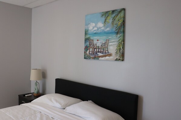 A bedroom with a bed, a nightstand with a lamp, and a painting of a beach scene on the wall.