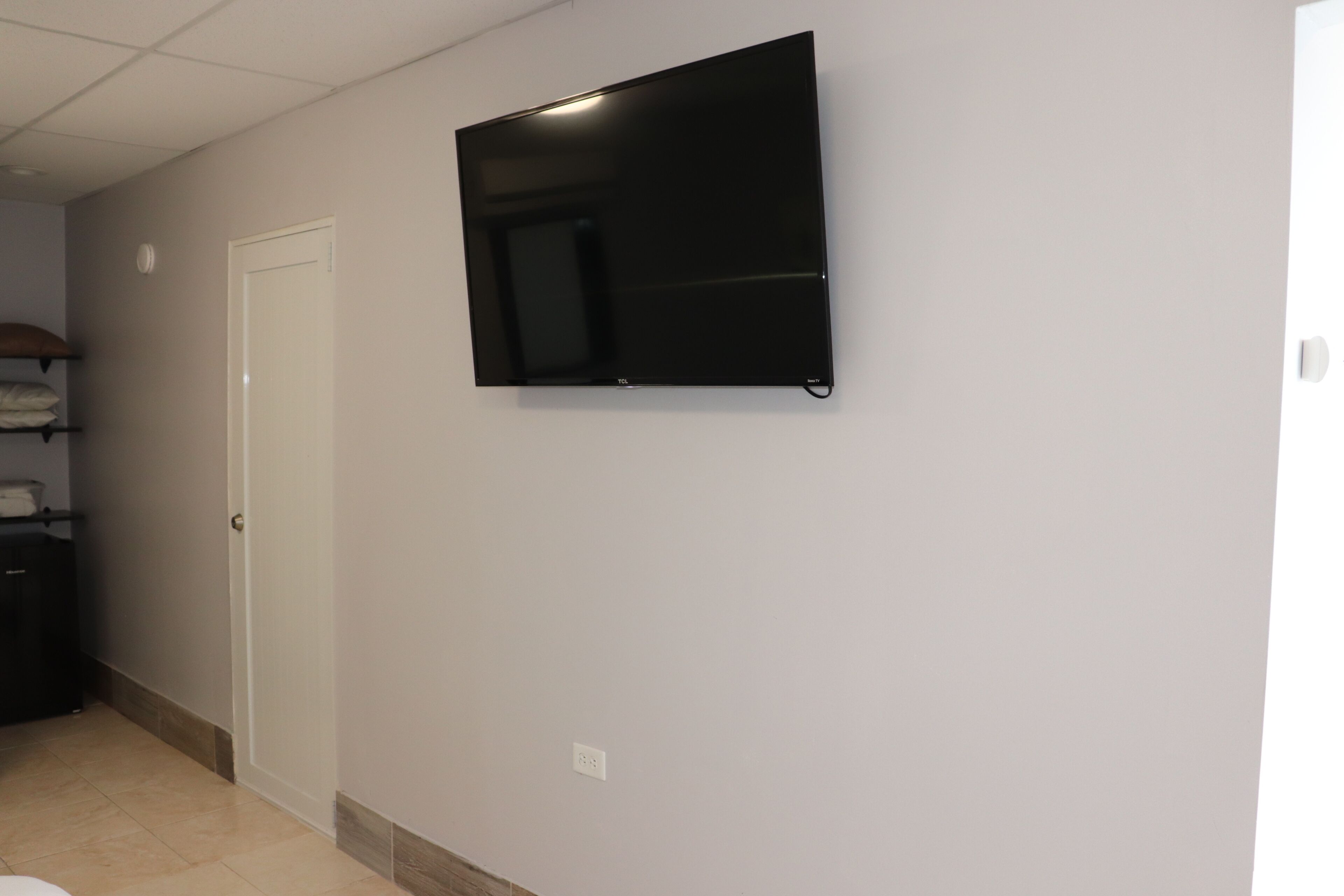 A room with a flat-screen TV mounted on the wall, a white door, and a tiled floor.