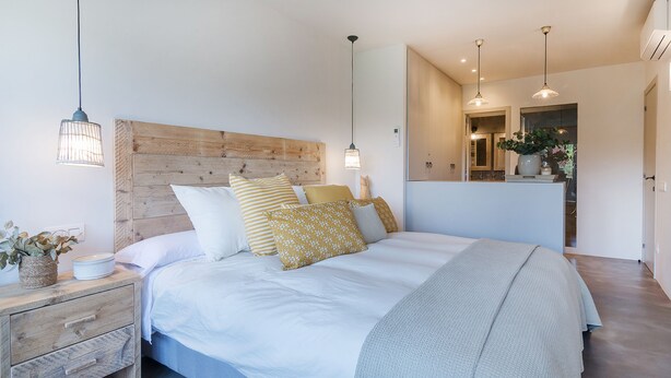 A modern bedroom with a wooden headboard, a large bed with white bedding, multiple pillows, bedside tables with lamps, and a view into the adjacent bathroom.