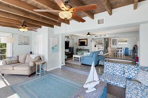 Relax in this spacious living area with a high ceiling and central fan, featuring a beige sectional sofa, a blue patterned sofa, and a small round table.