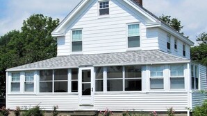 A two-story white house with a gabled roof and a chimney, surrounded by a well-maintained lawn and a stone border. - Sunny Jenness Beach View Home – Quiet Rd, Perfect for Families & Coastal Escapes (Rye)