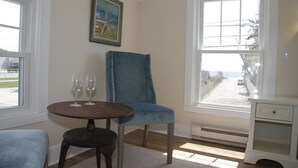 A room with a large window, a blue chair, a small table with two wine glasses, and a white nightstand. - Sunny Jenness Beach View Home – Quiet Rd, Perfect for Families & Coastal Escapes (Rye)