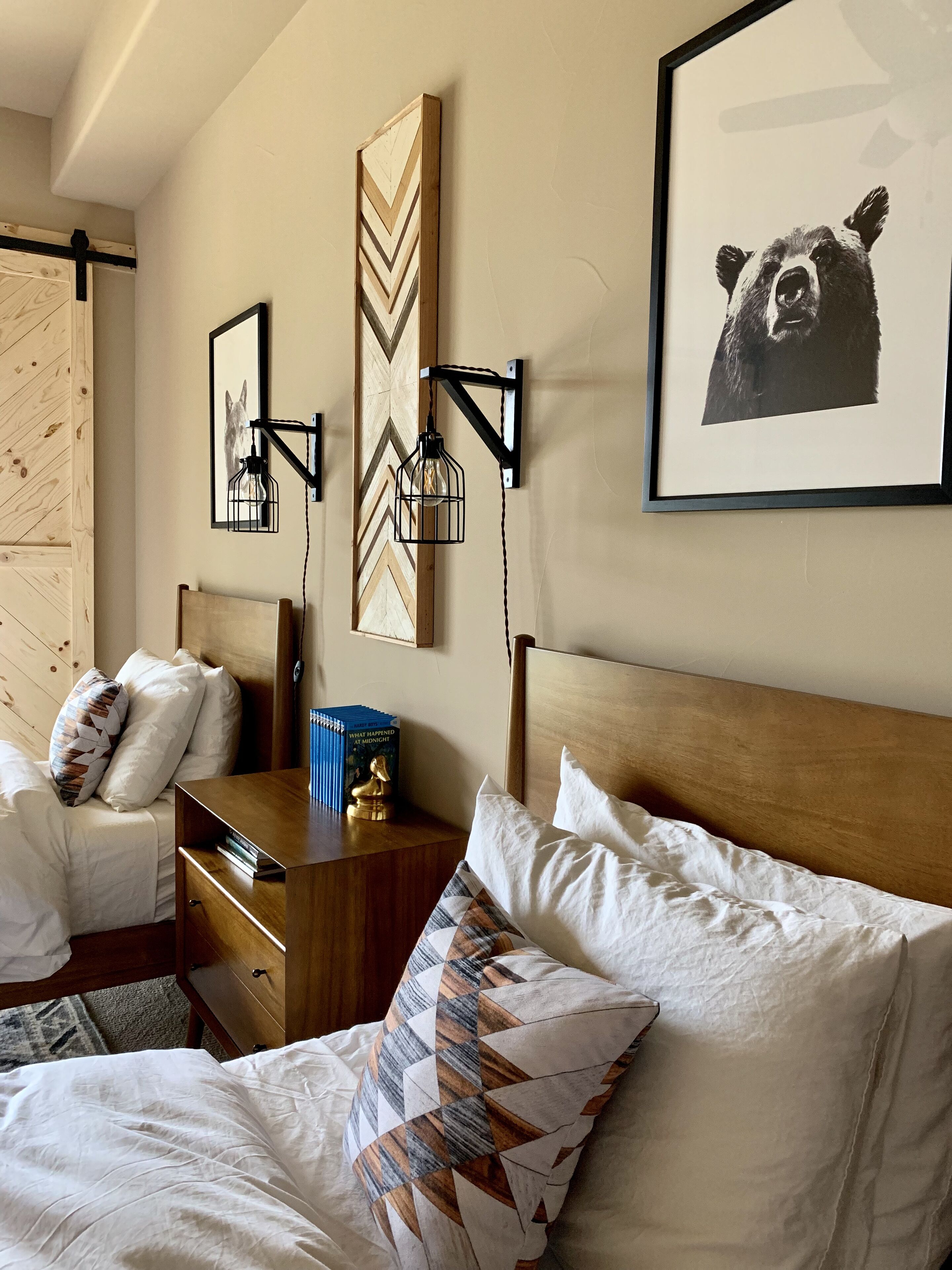 Une chambre d’hôtel avec une tête de lit en bois, un lit avec une literie blanche, une table de chevet avec un livre et un objet décoratif, un luminaire fixé au mur et un tableau représentant un ours.