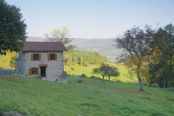 A stone house with a red-tiled roof, surrounded by a grassy field and trees.
