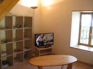 A room with a wooden bookshelf, a television on a stand, and a window with a view of greenery.