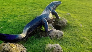 En stor skulptur af en leguan på en græsplæne med udsigt til en strand og bygninger i baggrunden.