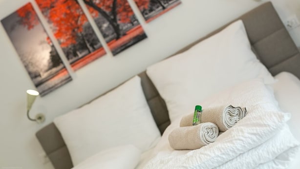 En pænt redt seng med sammenrullede håndklæder og en flaske lotion, en væg med tre billeder hængende, og en senget lampe.