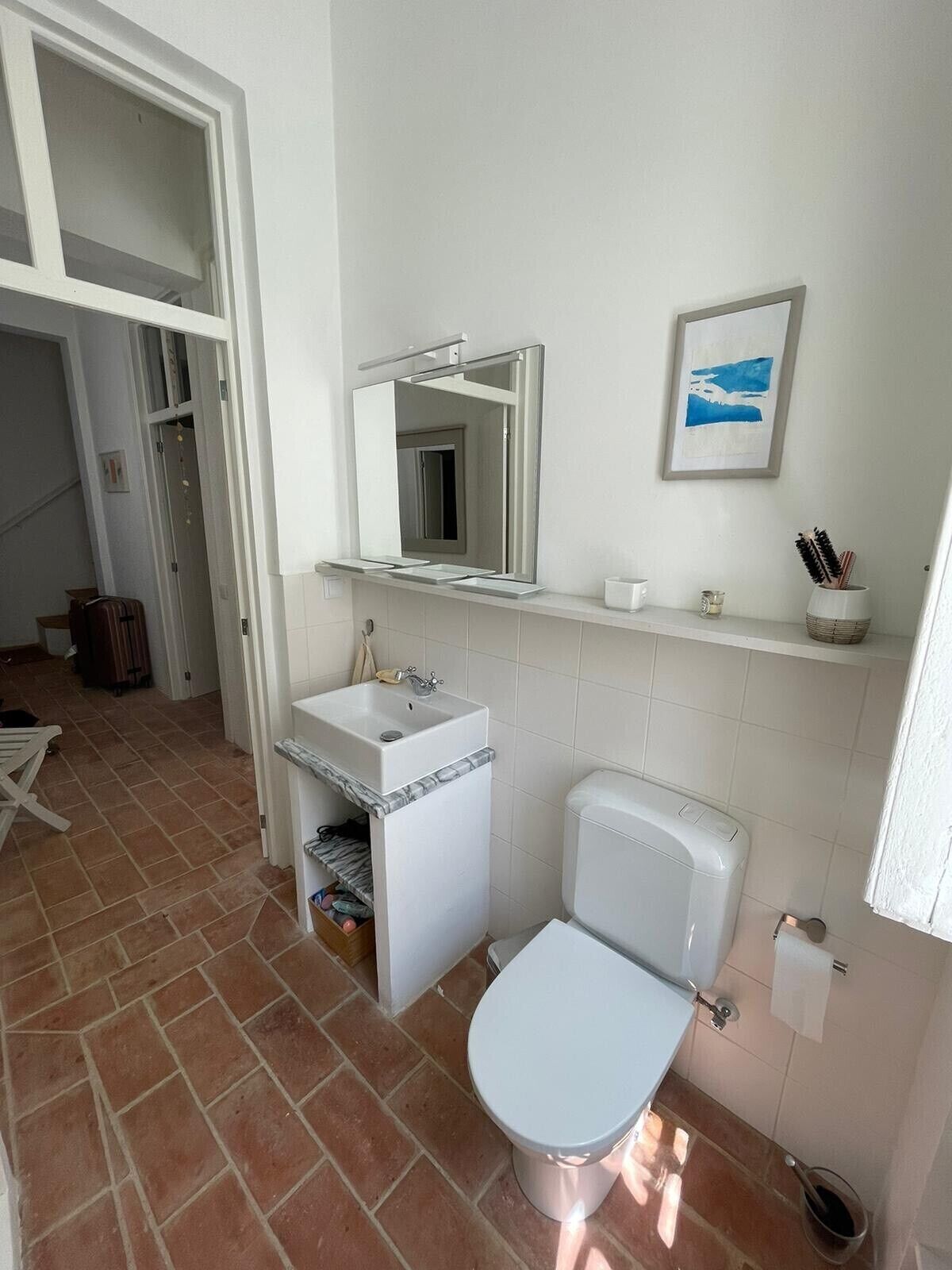 Une salle de bain avec un lavabo blanc, des toilettes et un miroir. On y trouve une étagère avec des articles de toilette et un tableau encadré accroché au mur.
