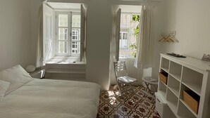 A bedroom with a bed, a desk, and a chair. There are two windows with curtains, and a white wall with a shelf.
