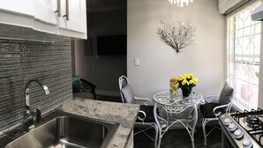 Une cuisine dotée d’un plan de travail en granit, d’un évier en acier inoxydable et d’une cuisinière à gaz. On y trouve une petite table ronde, sur laquelle est posé un vase rempli de fleurs jaunes, ainsi qu’une chaise.