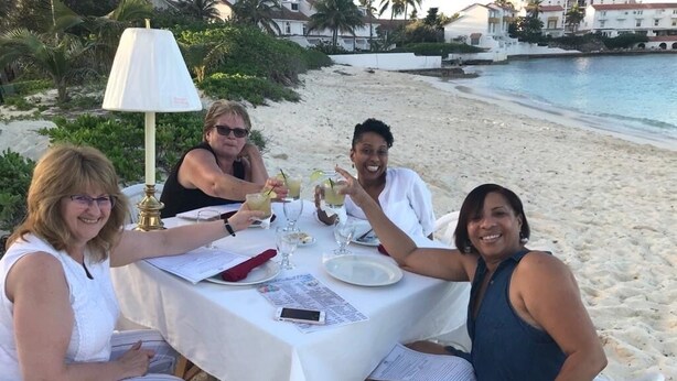 Un groupe de personnes est assis autour d’une table sur une plage, une nappe blanche recouvre la table, et elles savourent des boissons et des plats.