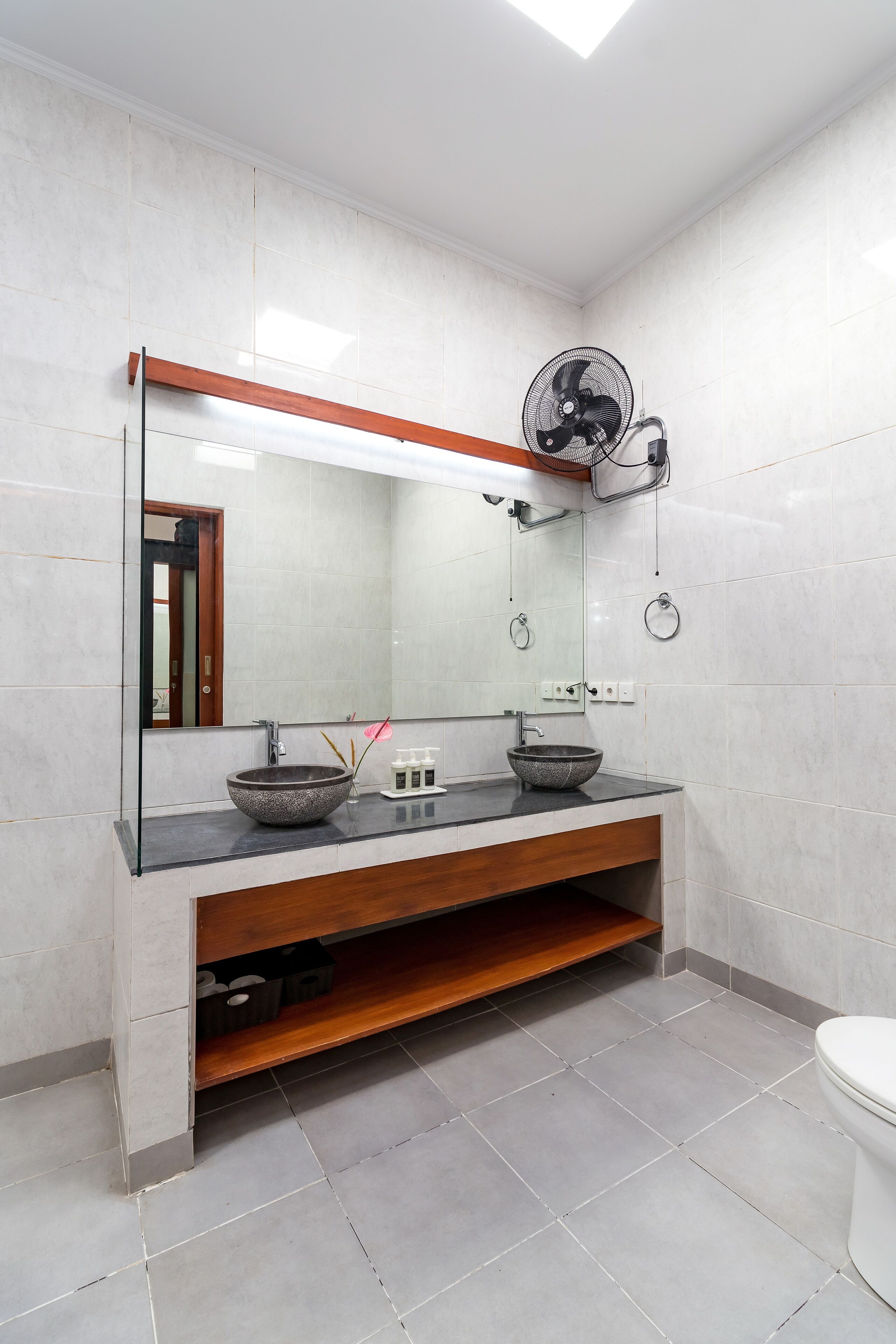 A bathroom with a large mirror, a vessel sink, a wooden shelf, a fan, and a toilet.
