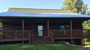 A two-story log cabin with a metal roof, a front porch with wooden railings, and a small front door.