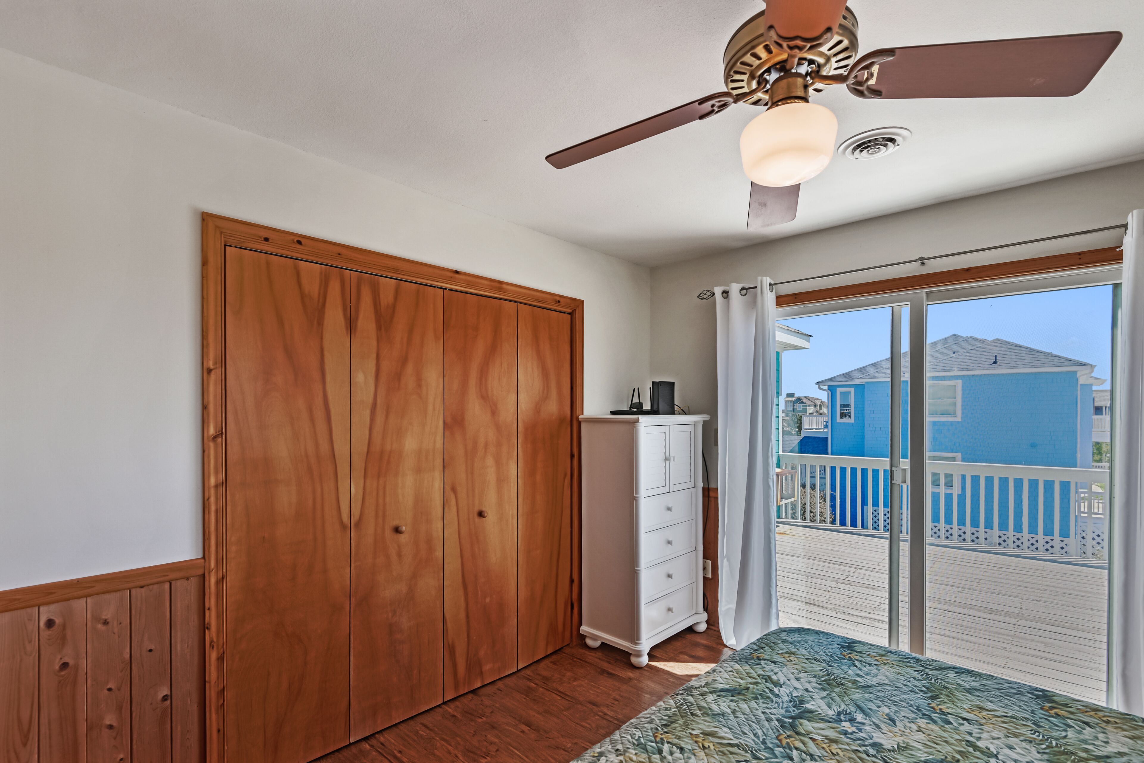 Une chambre avec une armoire en bois, une commode blanche, un ventilateur de plafond et une vue sur un immeuble bleu, visible à travers une porte-fenêtre coulissante.