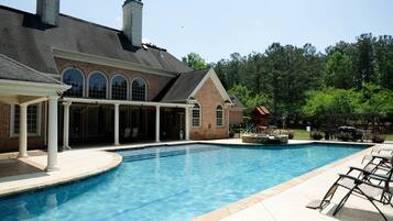 A swimming pool with a covered patio area, a brick building with arched windows, and a forested background.