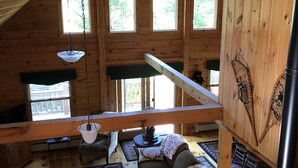 A cozy cabin with a loft bedroom, wooden walls, and large windows.