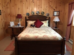 A wooden-paneled bedroom with a bed, two bedside tables, a lamp, a teddy bear, and decorative plates on the wall.