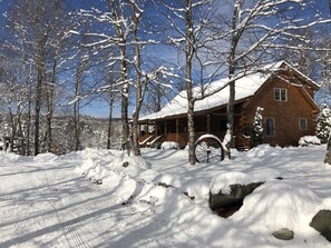 A wooden cabin with a porch, surrounded by snow-covered trees and a clear sky.