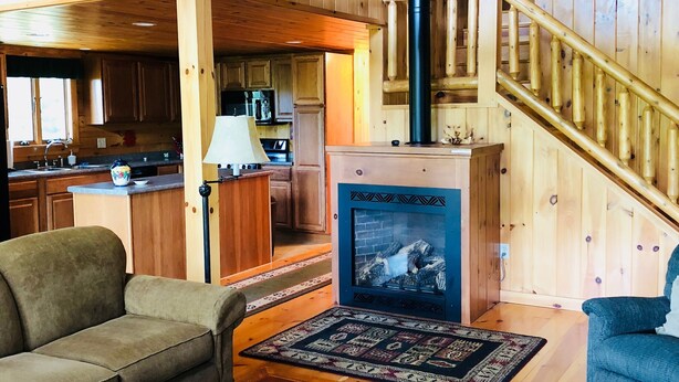 A cozy cabin interior with a wooden staircase, a fireplace, a sofa, a coffee table, and a kitchen area.