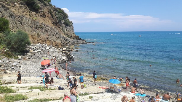 A rocky beach with clear blue water, people swimming and sunbathing, and a few boats in the distance. - Seaside cottage in residence (Costa Saracena)