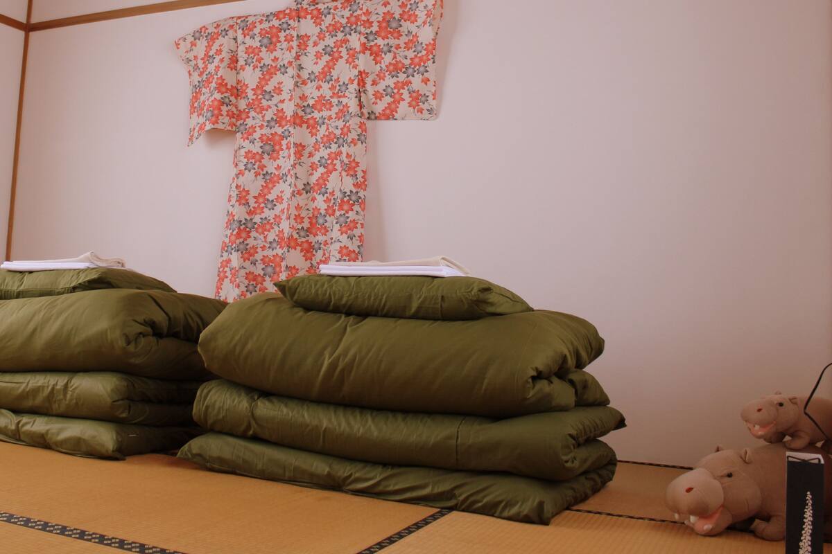 Una habitación con suelo de tatami, una cama futón con ropa de cama verde, un kimono floral colgado en la pared y un peluche de hipopótamo en el suelo.