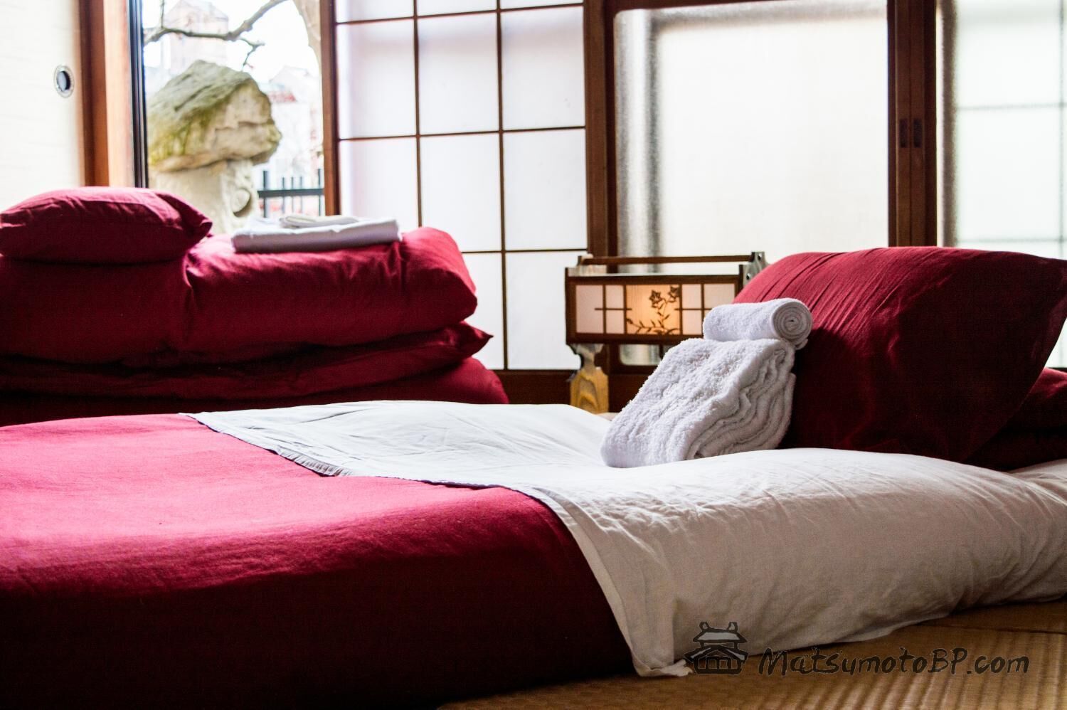 A hotel room with a bed, red bedding, a white pillow, and a nightstand with a lamp.